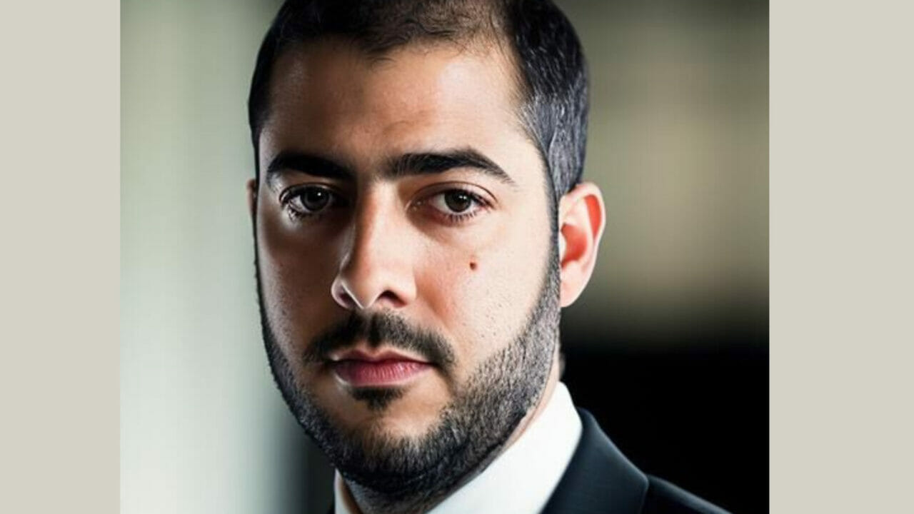 Foto de Guilherme. Homem de pele branca, cabelo curto preto e barba preta, olhos castanhos. Usa um terno preto, camisa branca e gravata azul escura.