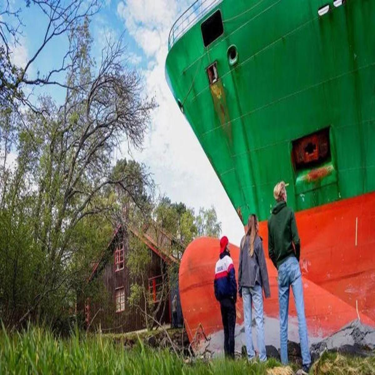 Navio encalhado próximo a casa de madeira observado por três pessoas em área arborizada