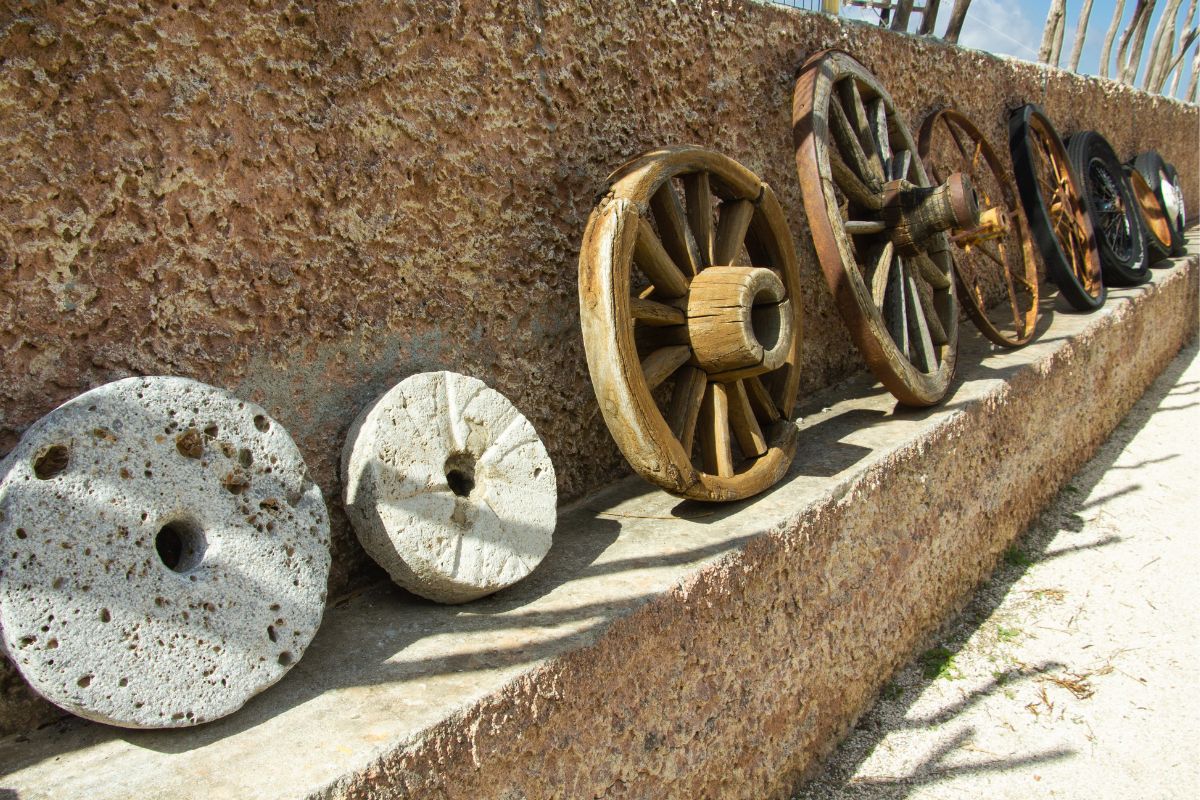 Várias rodas antigas de diferentes materiais como pedra, madeira e metal, dispostas lado a lado mostrando a evolução histórica das rodas.