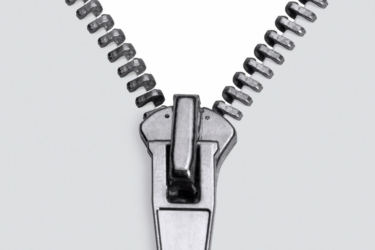 Zíper metálico prateado, detalhe em macro, com o cursor posicionado na parte inferior, mostrando os dentes se separando em um fundo branco e limpo. A imagem foca na textura e nos elementos mecânicos do fecho, transmitindo a ideia de abertura e funcionalidade.