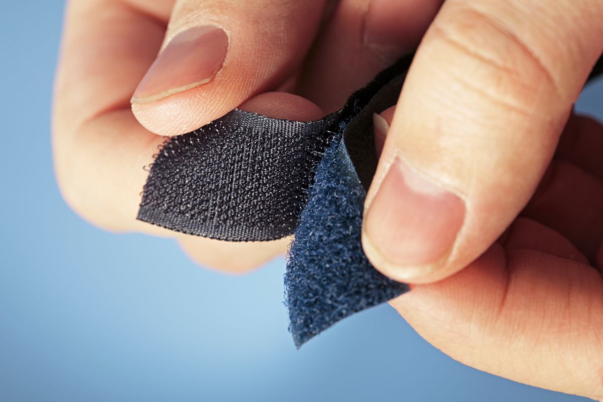 Close-up de dois dedos separando um fecho de velcro preto, mostrando os ganchos e laços intrincados.