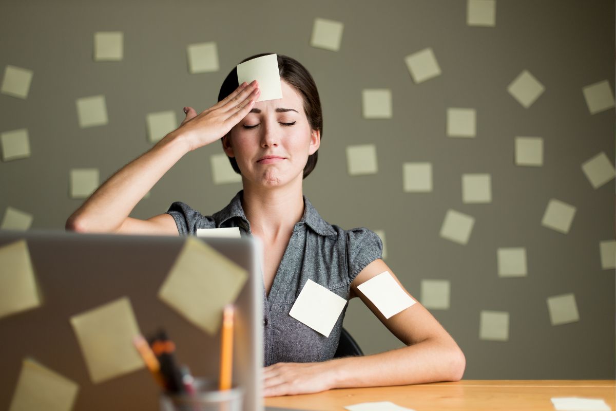 Mulher estressada com post-its no rosto e braço, sentada em frente a um laptop.