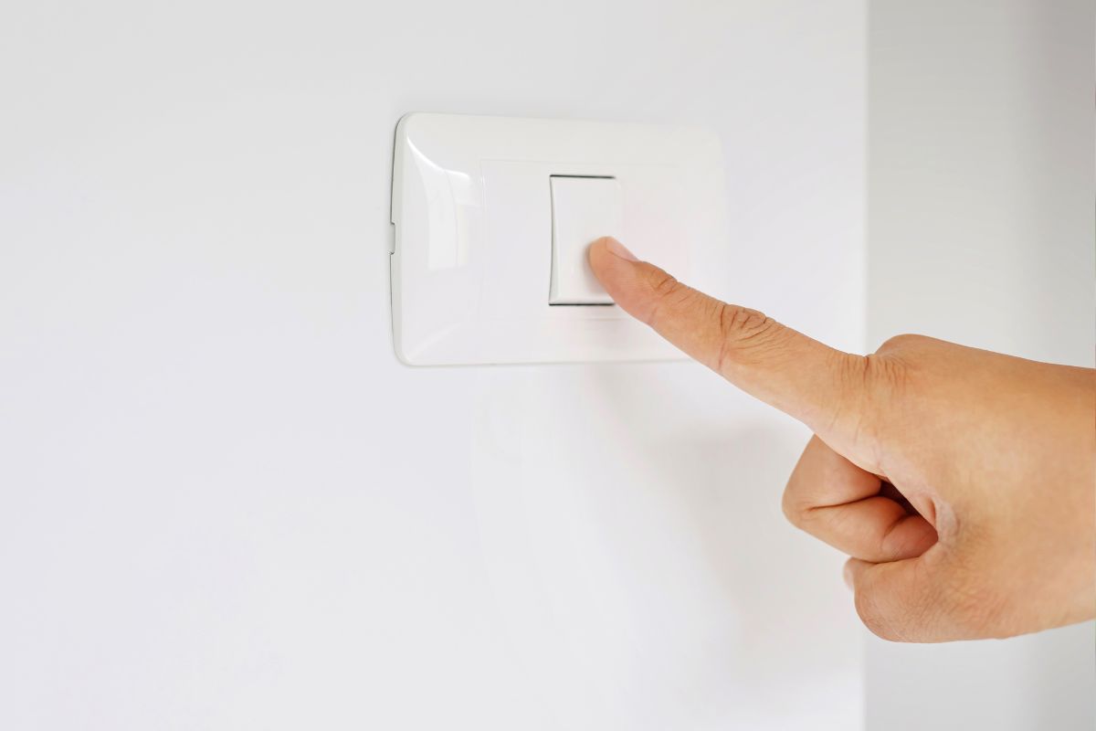 Mão de uma pessoa pressionando um interruptor de luz branco em uma parede branca.