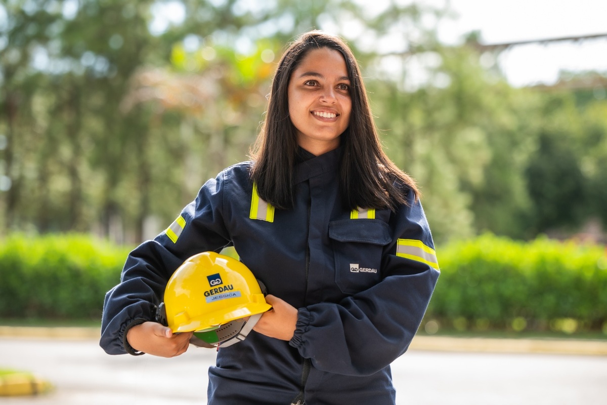 Mulher engenheira da Gerdau sorrindo, segurando capacete amarelo com uniforme de segurança.
