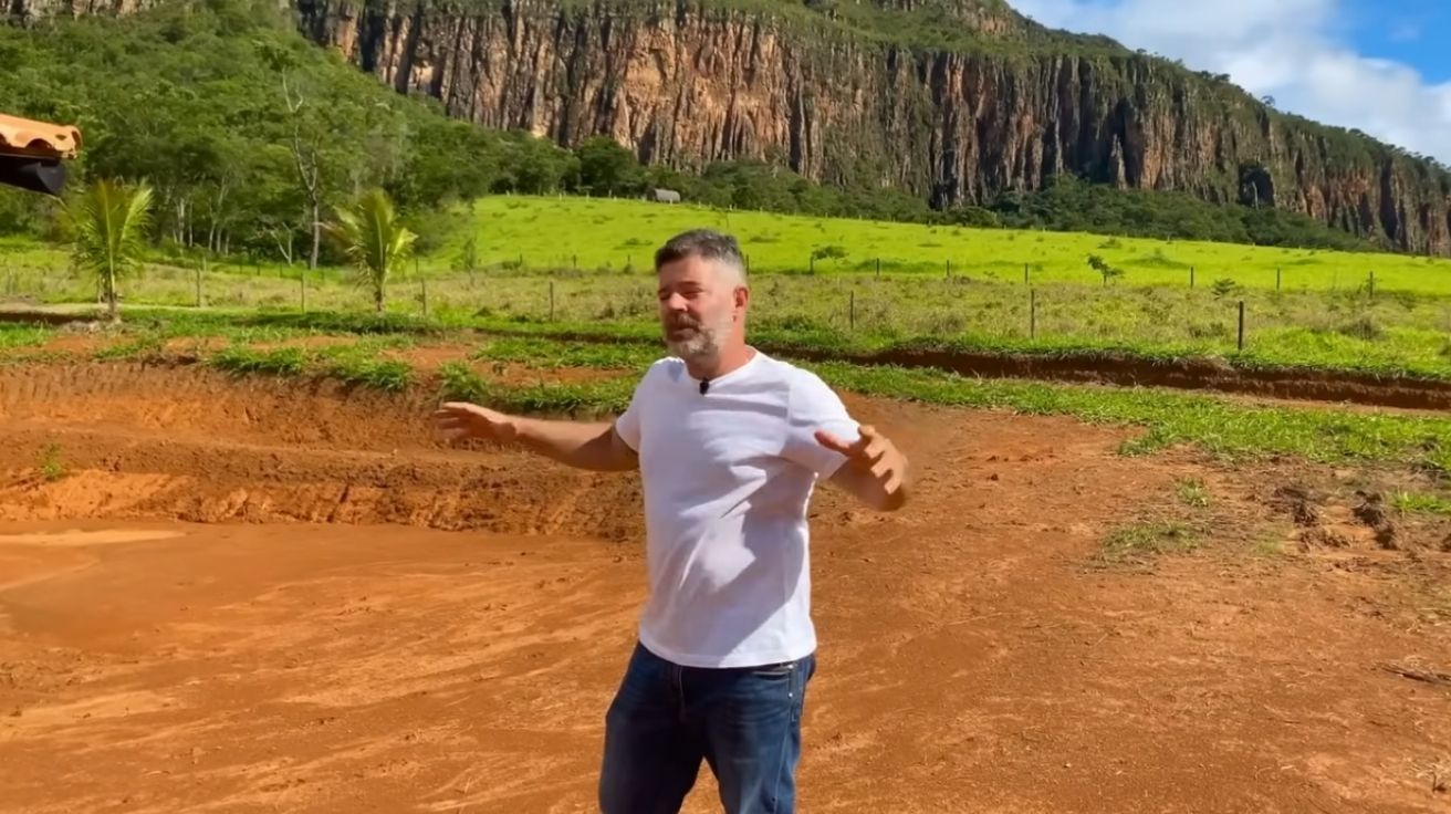 Homem em pé sobre solo vermelho em fazenda em Minas Gerais com paredão de pedra e vegetação ao fundo, em área de construção sustentável