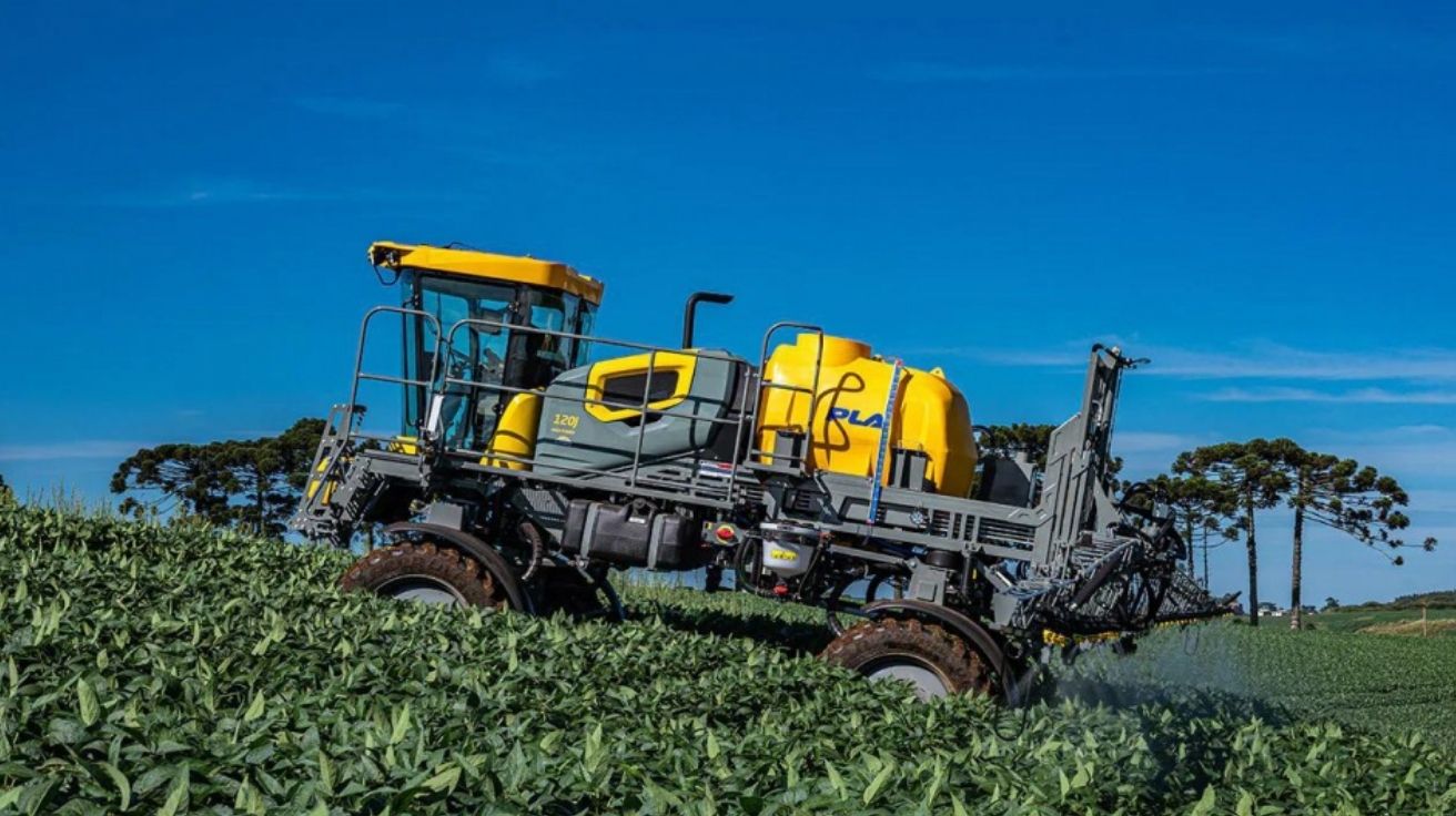 Pulverizador agrícola PLA 120J em lavoura de soja com barra de pulverização estendida e cabine panorâmica, operando em terreno inclinado.