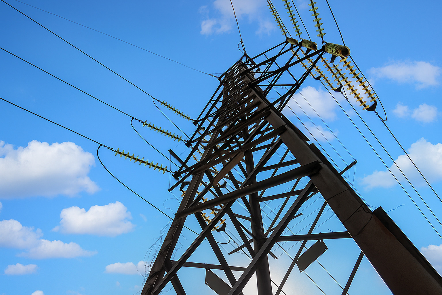 Vista de baixo para cima de uma torre de transmissão de energia elétrica contra um céu azul com poucas nuvens espalhadas.
