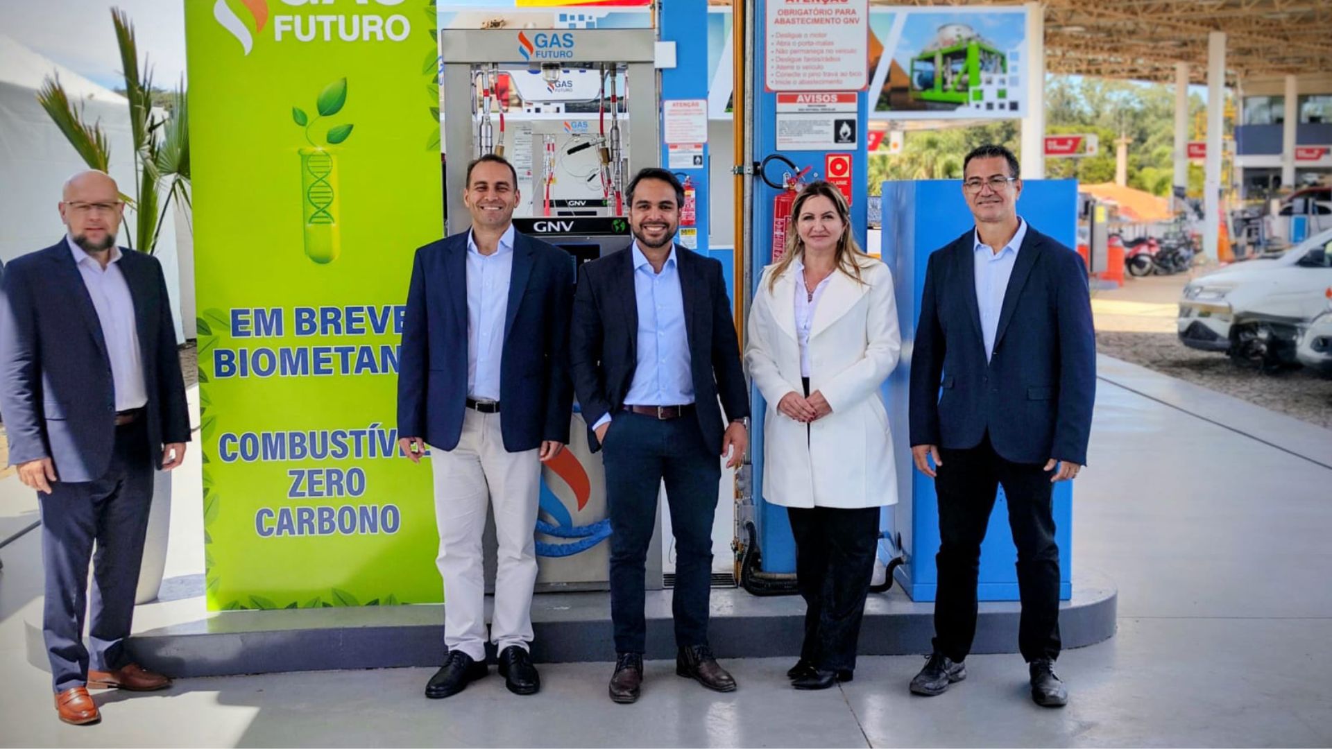 Cinco pessoas em traje formal posam em frente a um posto de abastecimento com gás natural veicular e um painel verde que anuncia a chegada do biometano como combustível zero carbono.