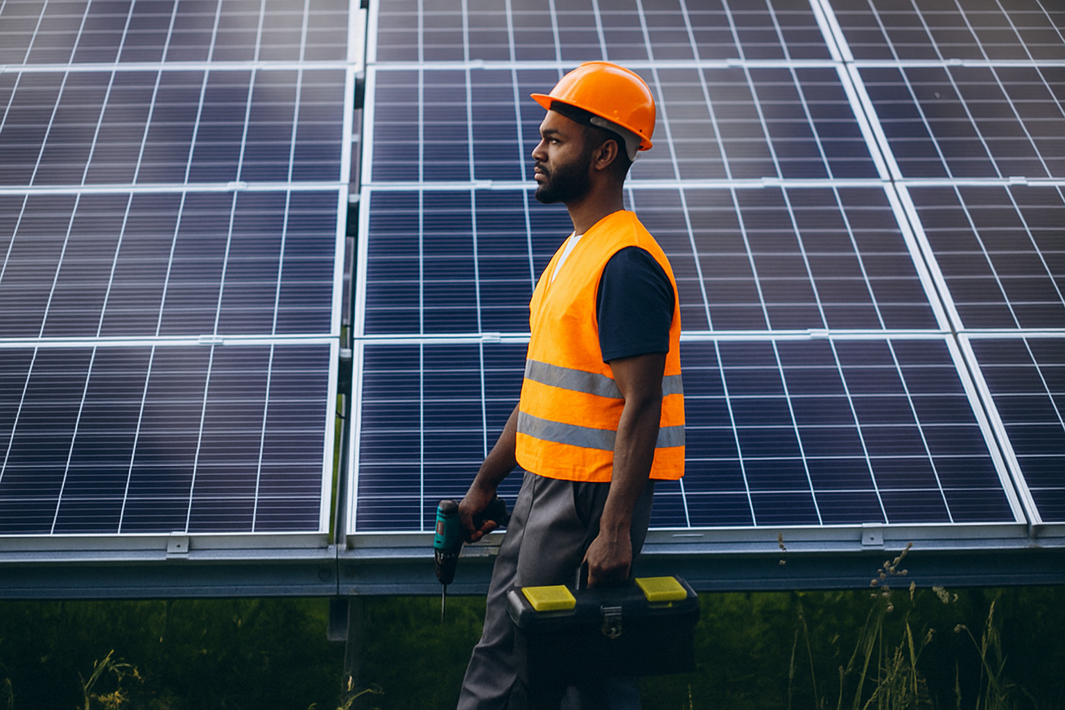 Trabalhador negro com uniforme de segurança caminhando em frente a painéis solares.