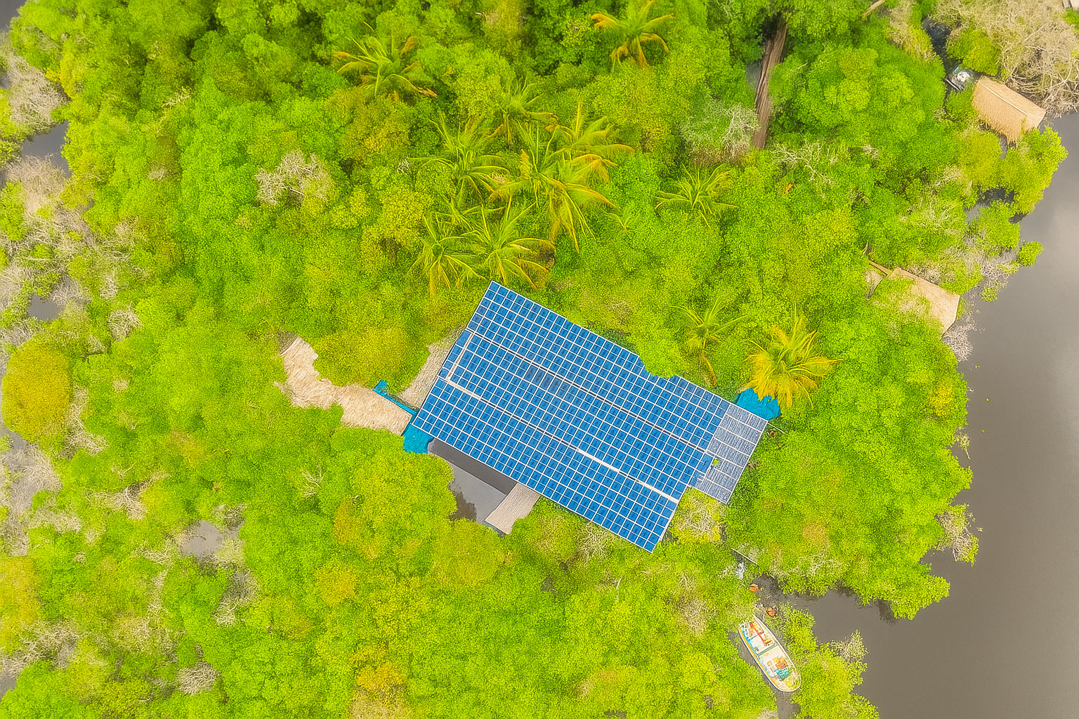Vista aérea de uma construção com telhado coberto por painéis solares no meio da floresta amazônica ao meio-dia.