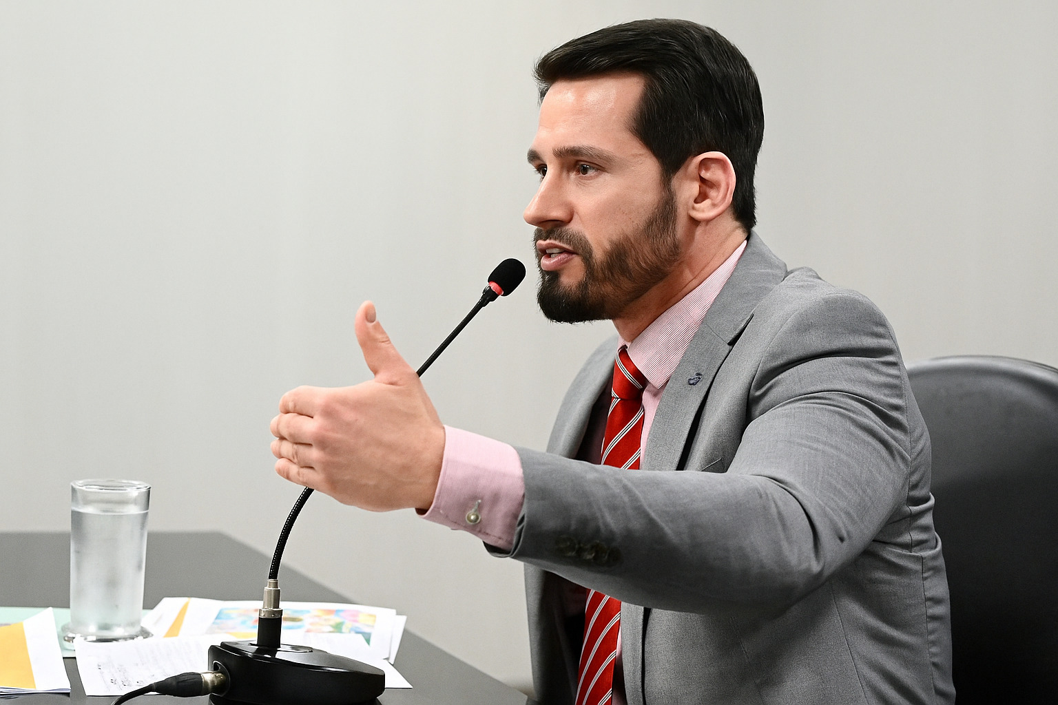 Homem de terno cinza e gravata vermelha fala ao microfone durante reunião oficial.