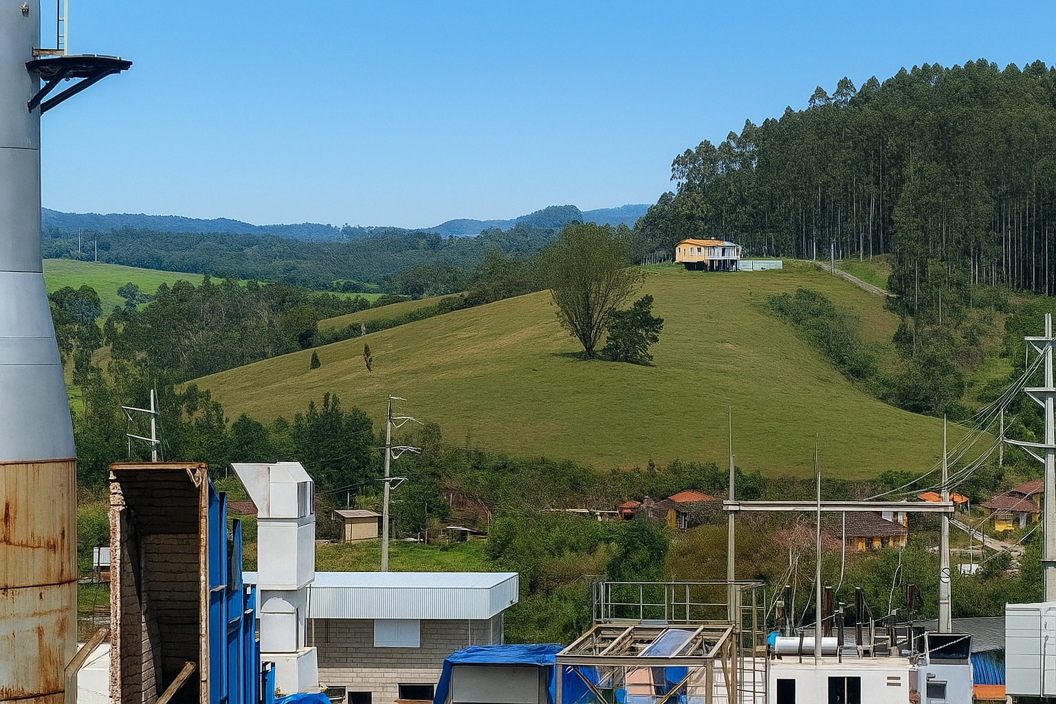 Vista de uma usina de energia com chaminé e estruturas industriais em frente a colinas verdes sob céu parcialmente limpo.