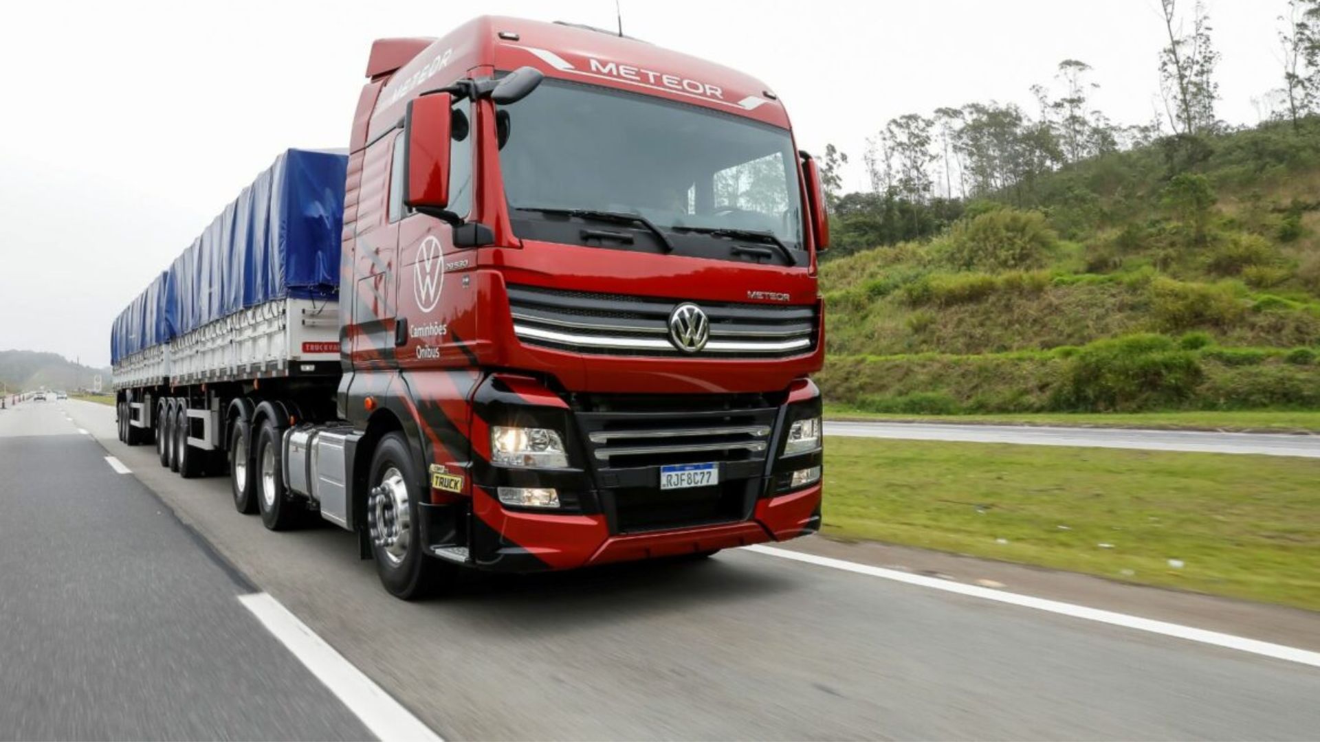 Caminhão Volkswagen Meteor vermelho com reboque coberto por lona azul trafegando em rodovia, representando transporte rodoviário de carga pesada.