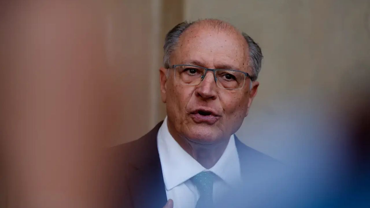 Geraldo Alckmin usando terno escuro e gravata verde, fala em público com expressão engajada, em ambiente formal.