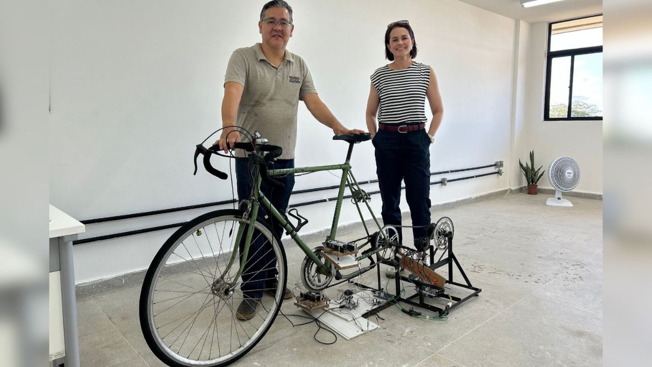 Dois pesquisadores ao lado de uma bicicleta estacionária modificada, conectada a um sistema mecânico e eletrônico para geração de energia limpa, em ambiente de laboratório.
