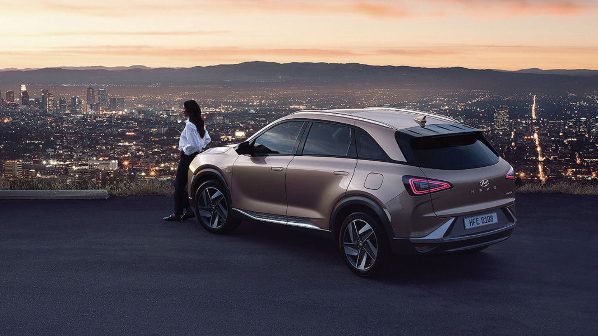 Pessoa ao lado de um Hyundai NEXO bronze estacionado em área elevada com vista para cidade ao pôr do sol.