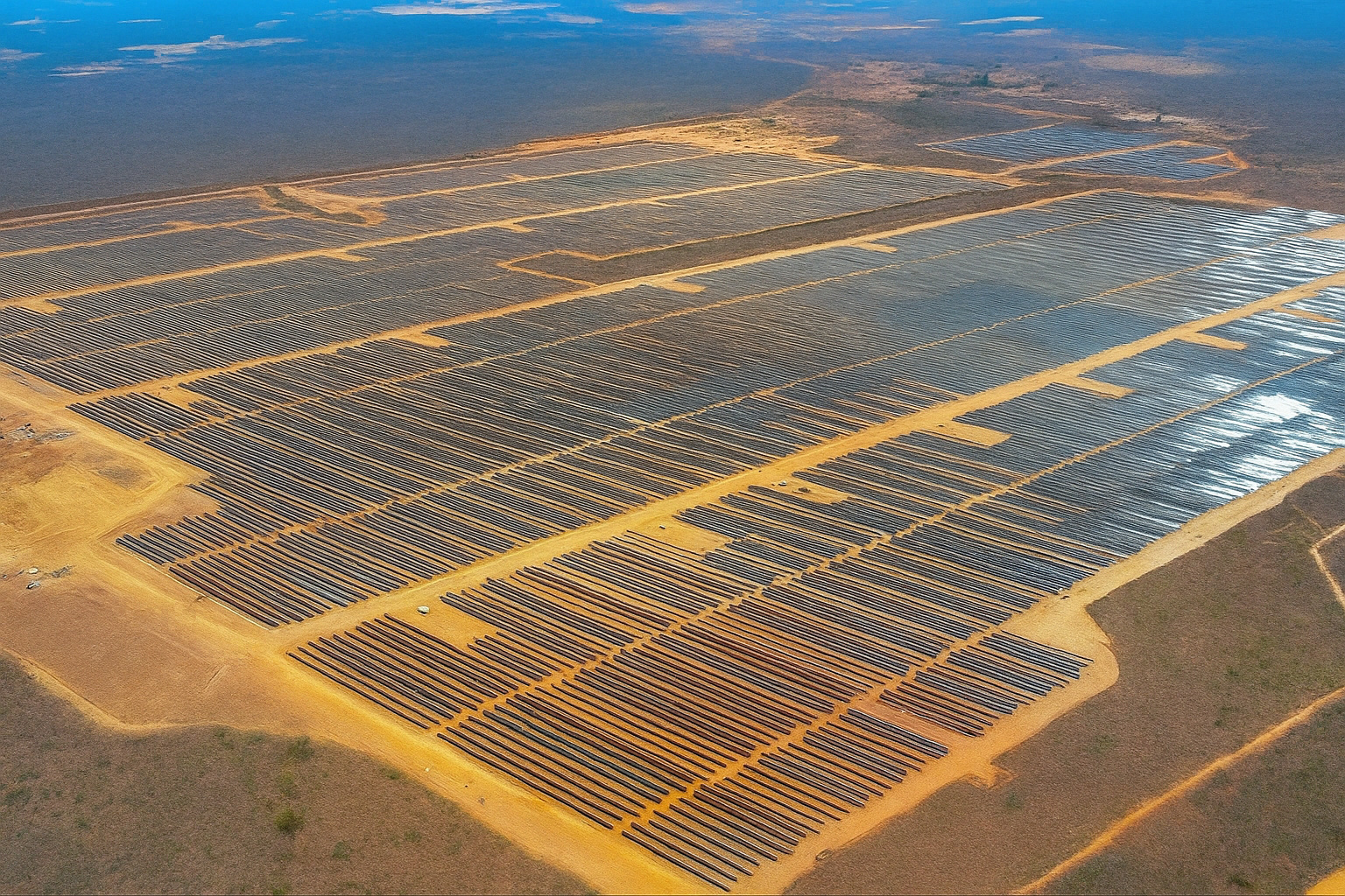 Vista aérea de uma usina solar em uma grande área de terreno seco, com painéis solares organizados em longas fileiras.