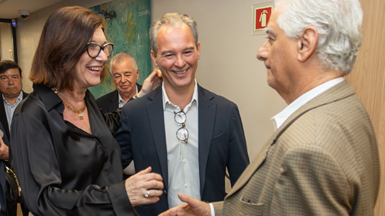 Magda Chambriard, Luiz Césio Caetano e Emiliano Gomes interagem cordialmente em evento profissional, trocando sorrisos e gestos amistosos em ambiente interno bem iluminado.