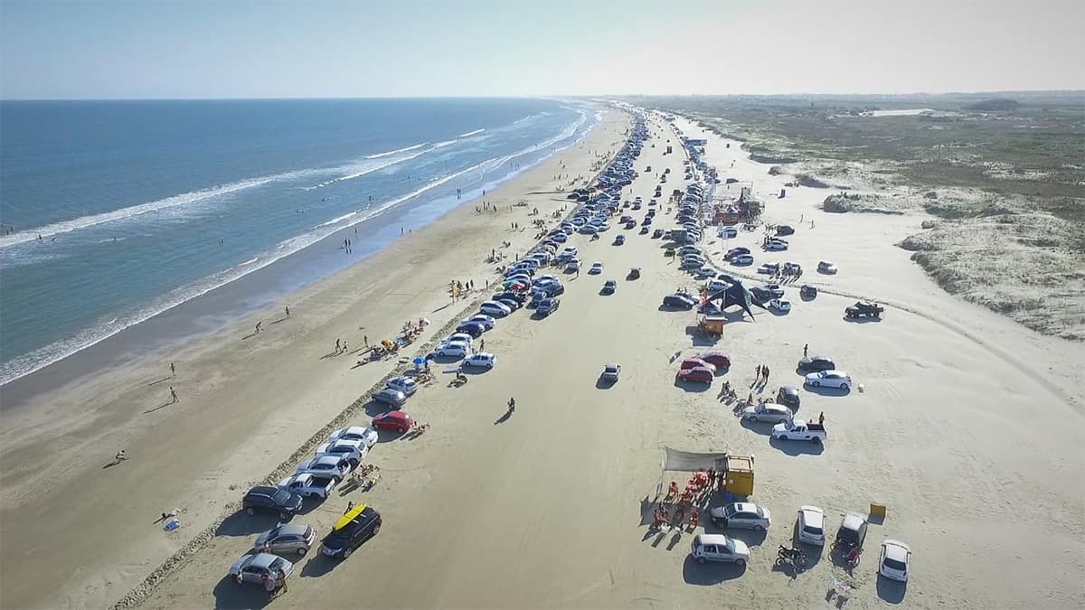 Vista aérea da Praia do Cassino, no Rio Grande do Sul, mostrando carros estacionados na faixa de areia e banhistas ao longo dos 254 km de extensão.