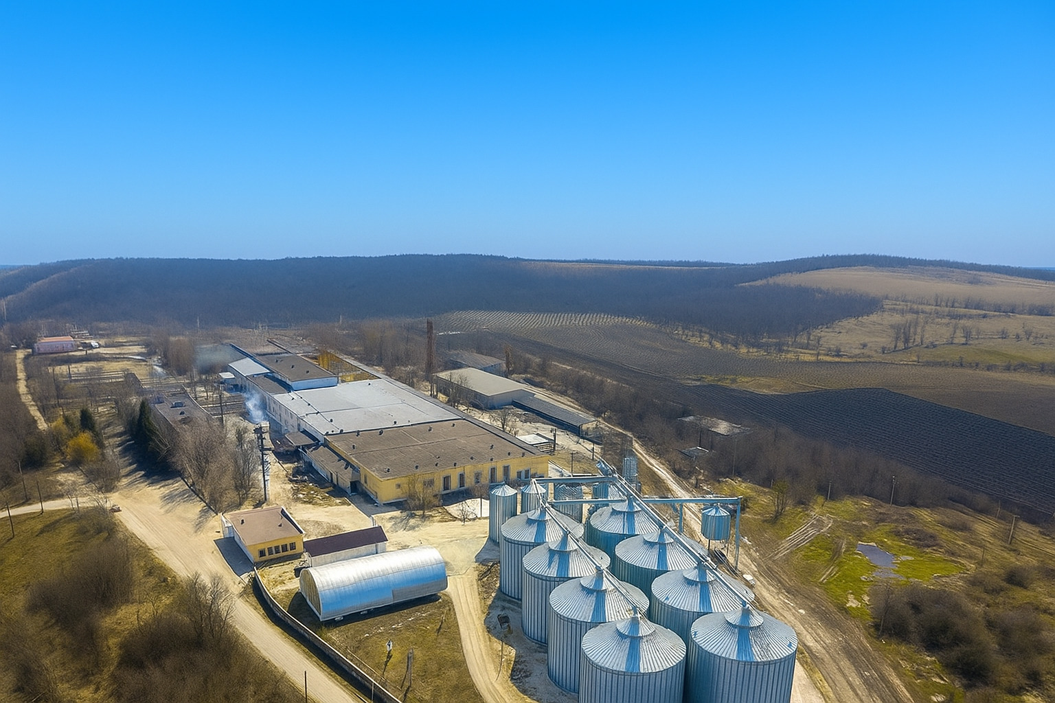 Vista aérea de uma indústria com silos metálicos e galpões sob céu claro ao meio-dia.