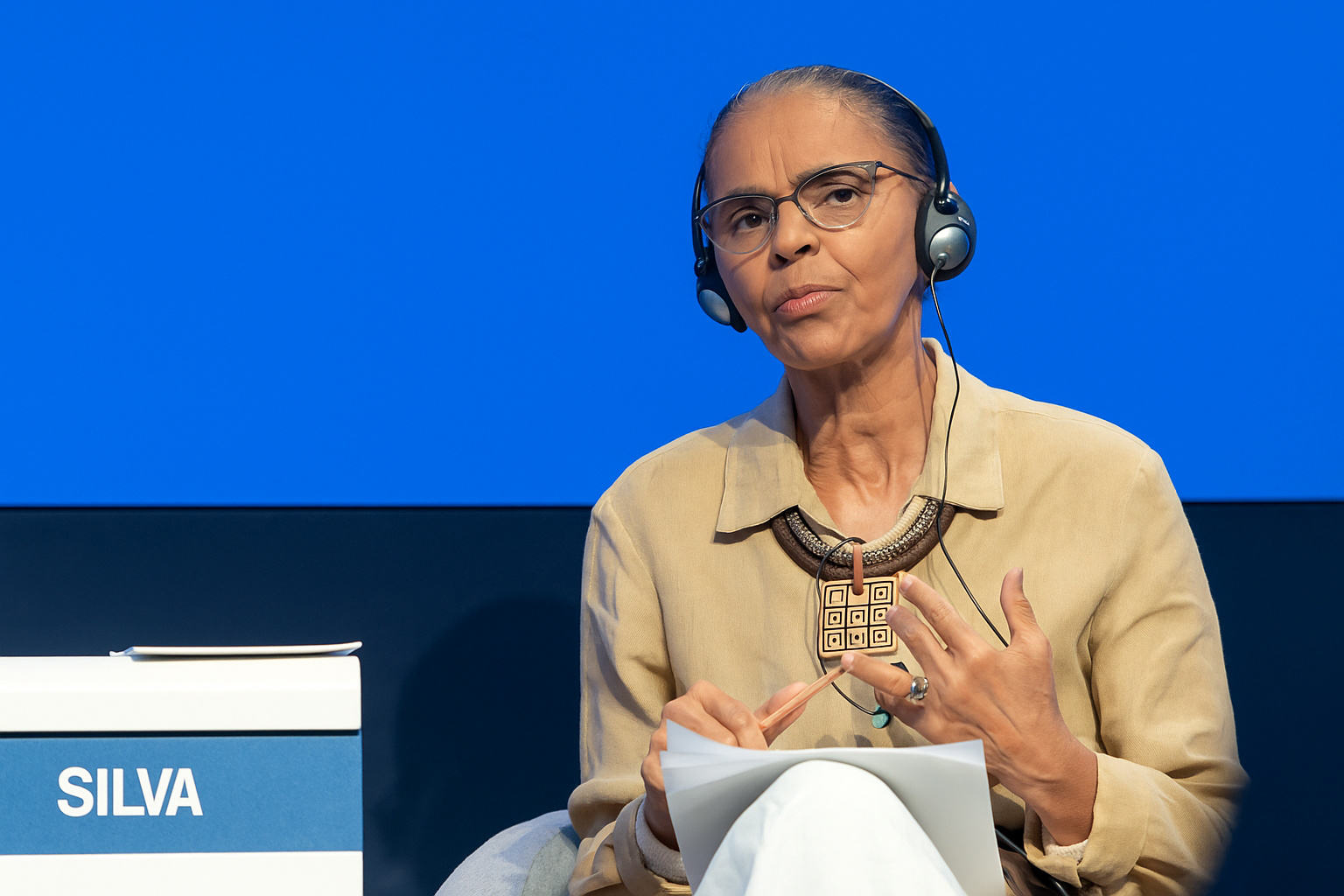 Mulher usando fones de ouvido e óculos participa de um painel de discussão diante de um fundo azul sólido.