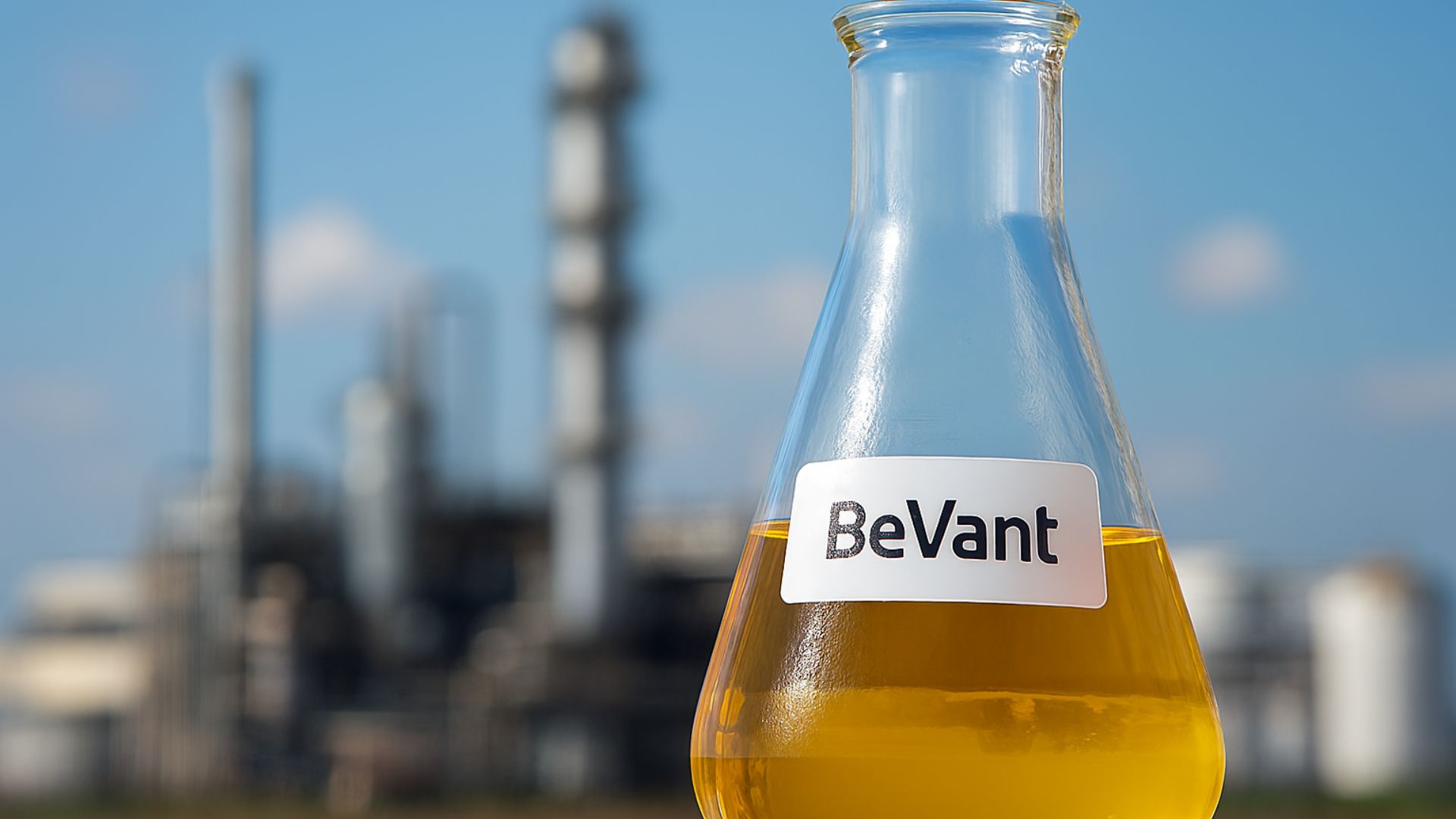 Frasco Erlenmeyer com biocombustível BeVant em destaque, com usina de bioenergia desfocada ao fundo sob céu azul