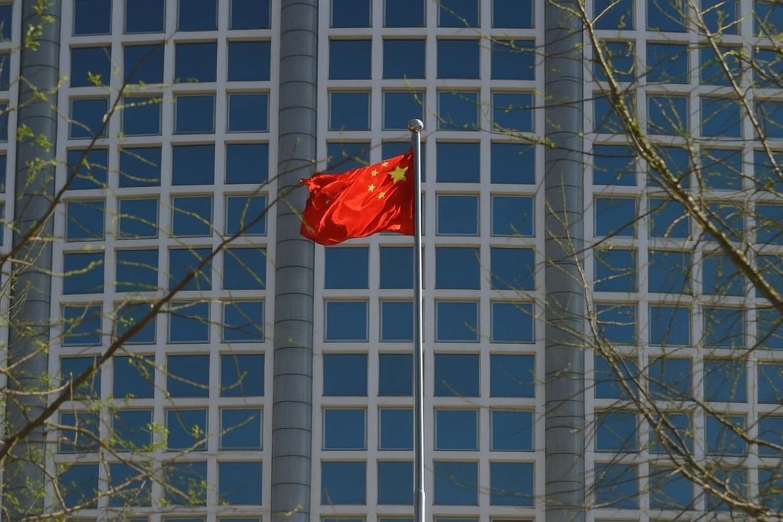 Bandeira da China tremulando em frente a um prédio envidraçado com poucas folhas nas árvores ao redor.