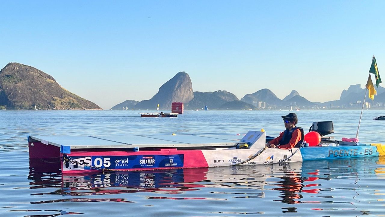 Pessoa conduzindo barco solar em competição, com painéis fotovoltaicos e paisagem urbana ao fundo