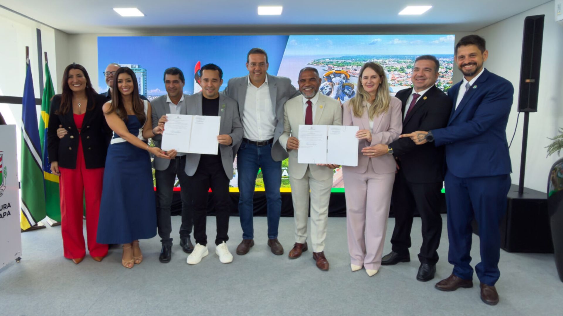 Grupo de autoridades e representantes reunidos em um evento oficial, posando com documentos assinados diante de um painel digital e bandeiras ao fundo.