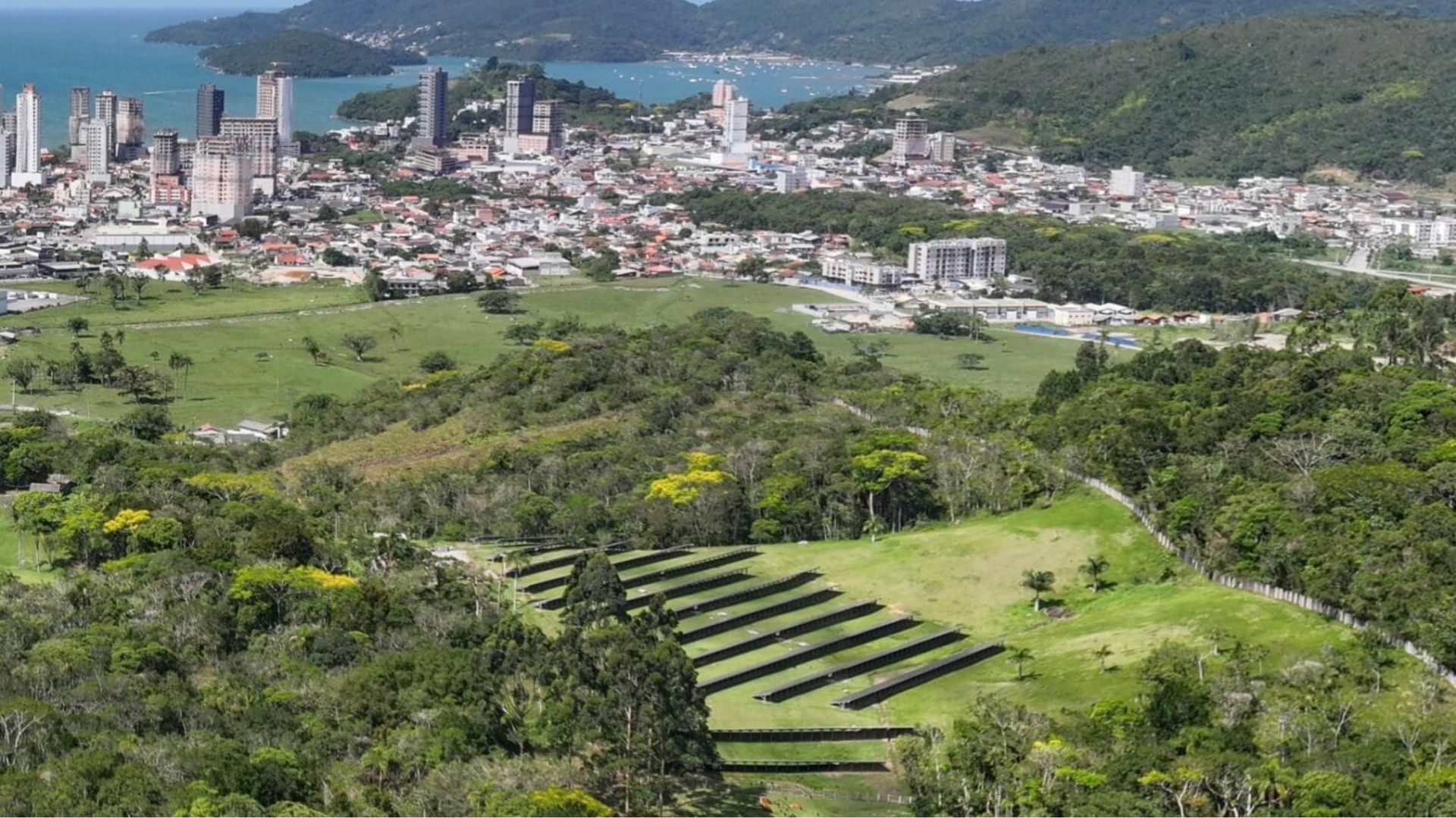 Fazenda solar instalada em área verde de Santa Catarina com painéis fotovoltaicos ao fundo e cidade litorânea ao redor, destacando geração de energia solar e preservação ambiental.