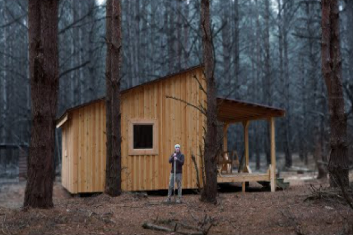 Cabana de madeira construída manualmente na floresta com morador em frente à estrutura artesanal.