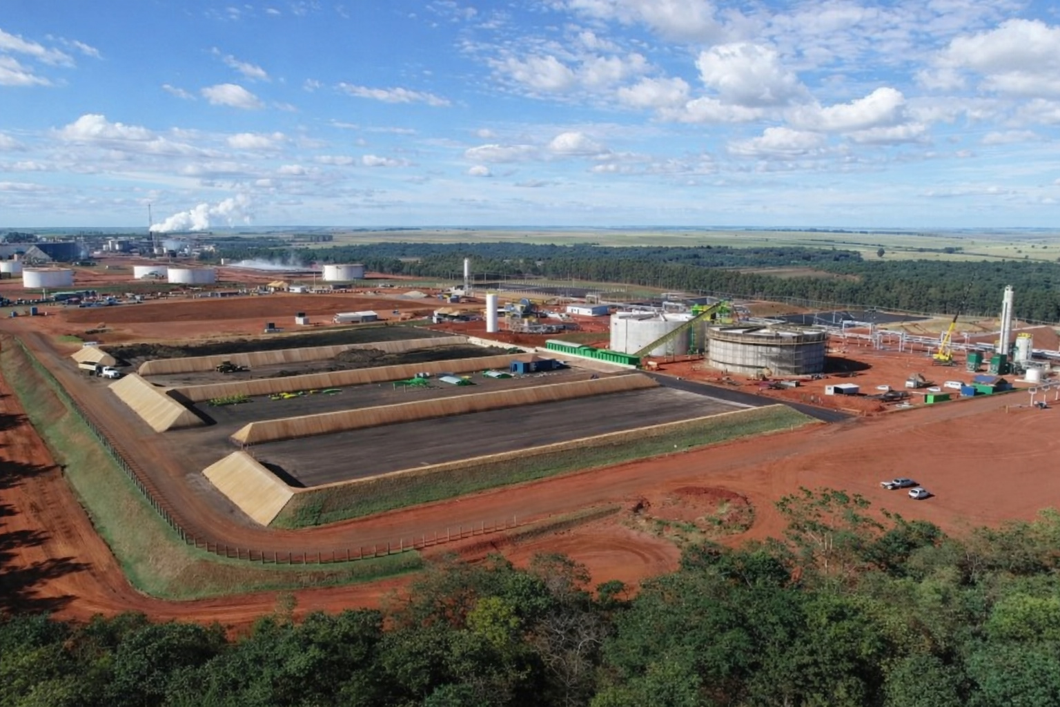 Vista aérea de complexo industrial com tanques, estruturas metálicas e áreas de armazenamento, cercado por vegetação, sob céu azul com algumas nuvens.