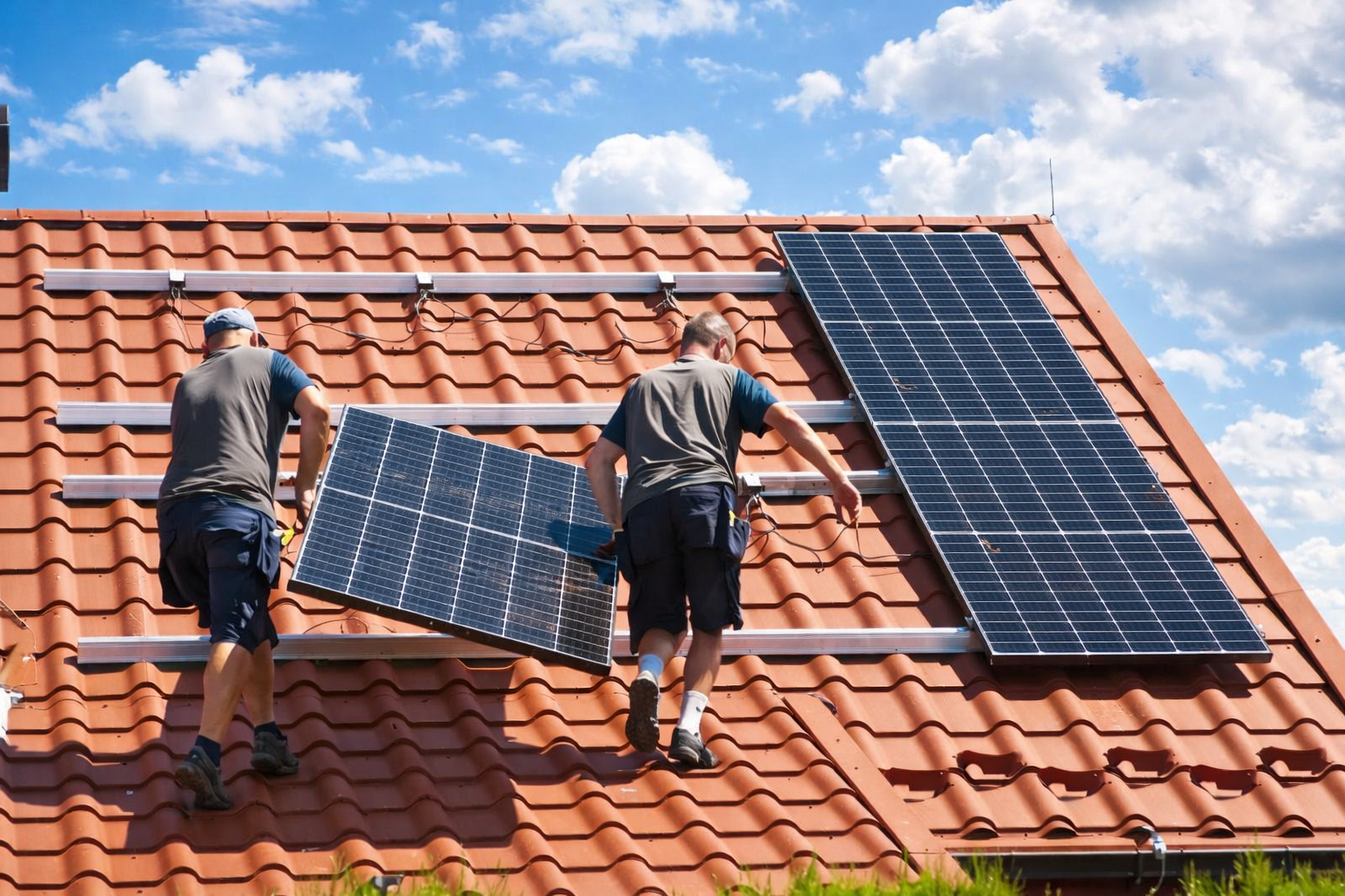 Dois técnicos instalando painéis solares em um telhado de telhas cerâmicas sob um céu azul com nuvens brancas.