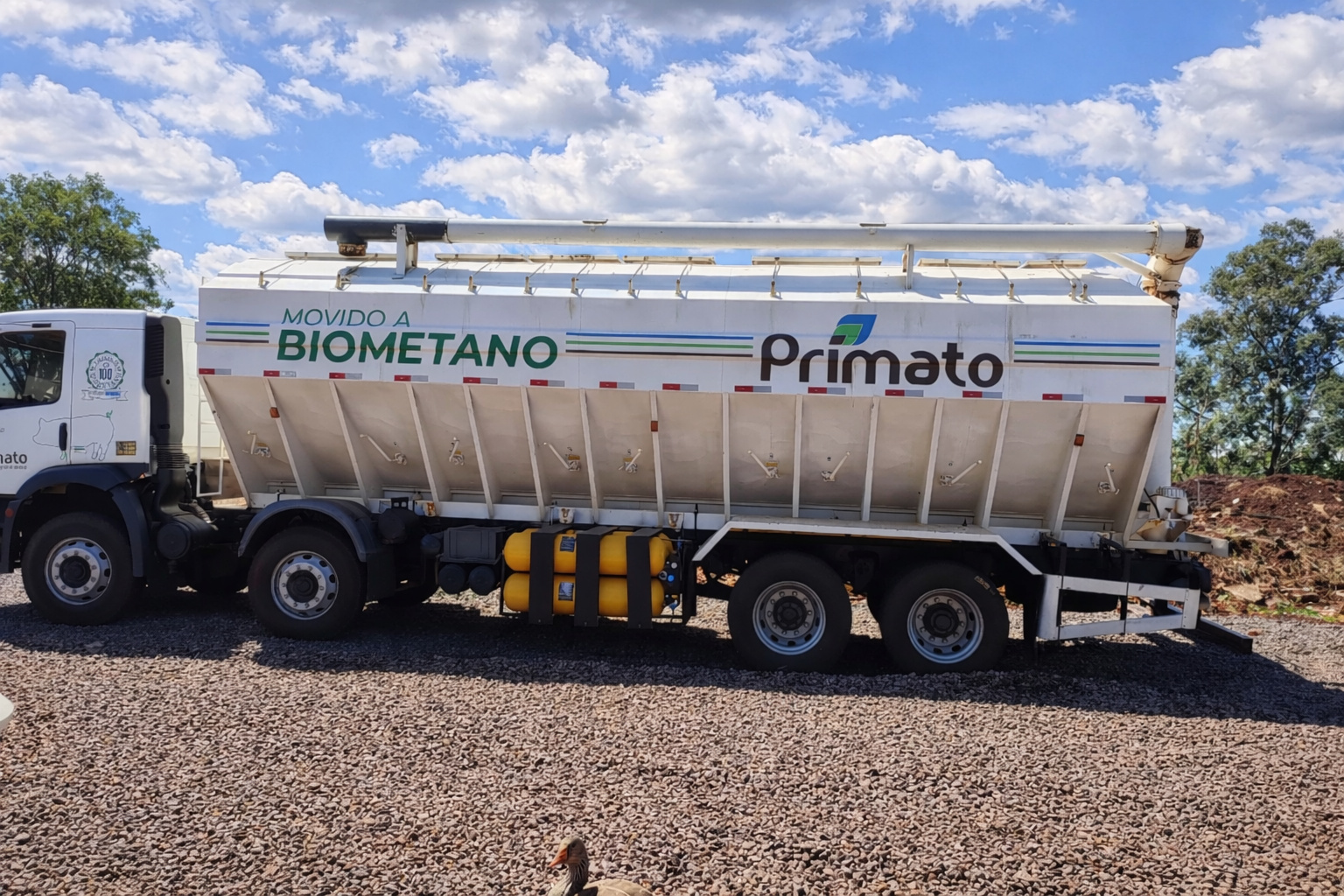 Caminhão-tanque movido a biometano estacionado em área rural sob céu com nuvens, com árvores e montes de terra ao fundo.