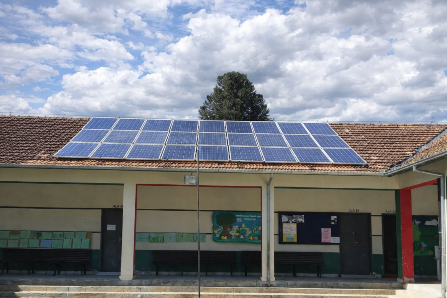 Prédio escolar de um andar com painéis solares no telhado, pátio com bancos e céu parcialmente nublado ao fundo.