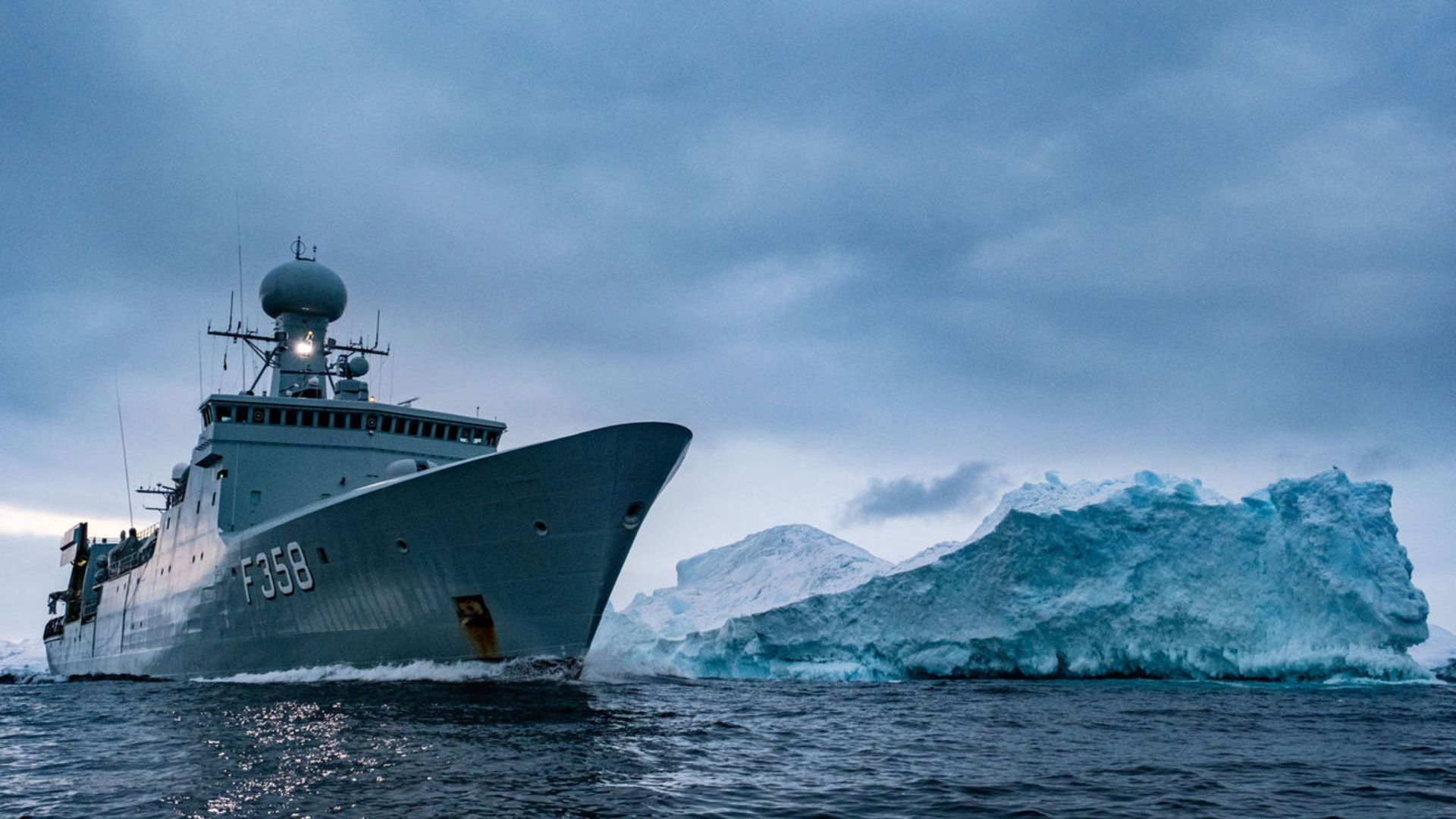Fragata F358 da Marinha Real Dinamarquesa navegando em águas geladas próximas a um iceberg, representando a presença militar no Ártico diante da disputa por petróleo e gás.