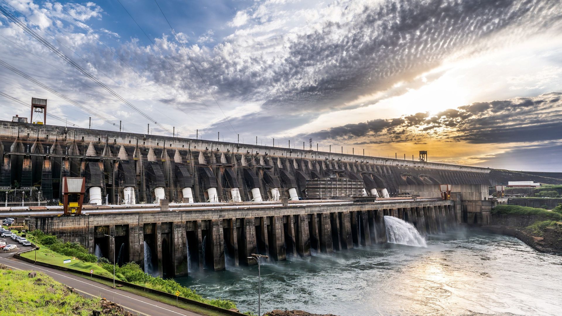Vista panorâmica da Usina Hidrelétrica de Itaipu, com vertedouro liberando água e estrutura monumental da barragem no Rio Paraná, ao entardecer.
