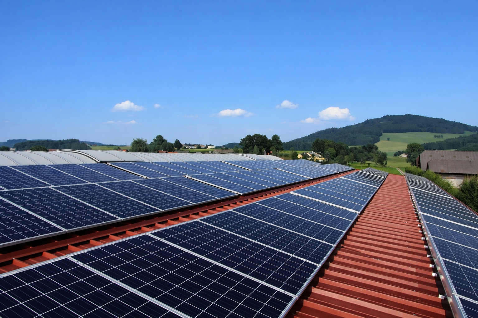 Painéis solares instalados em telhado metálico vermelho em área rural, com colinas verdes e céu azul com poucas nuvens ao meio-dia.