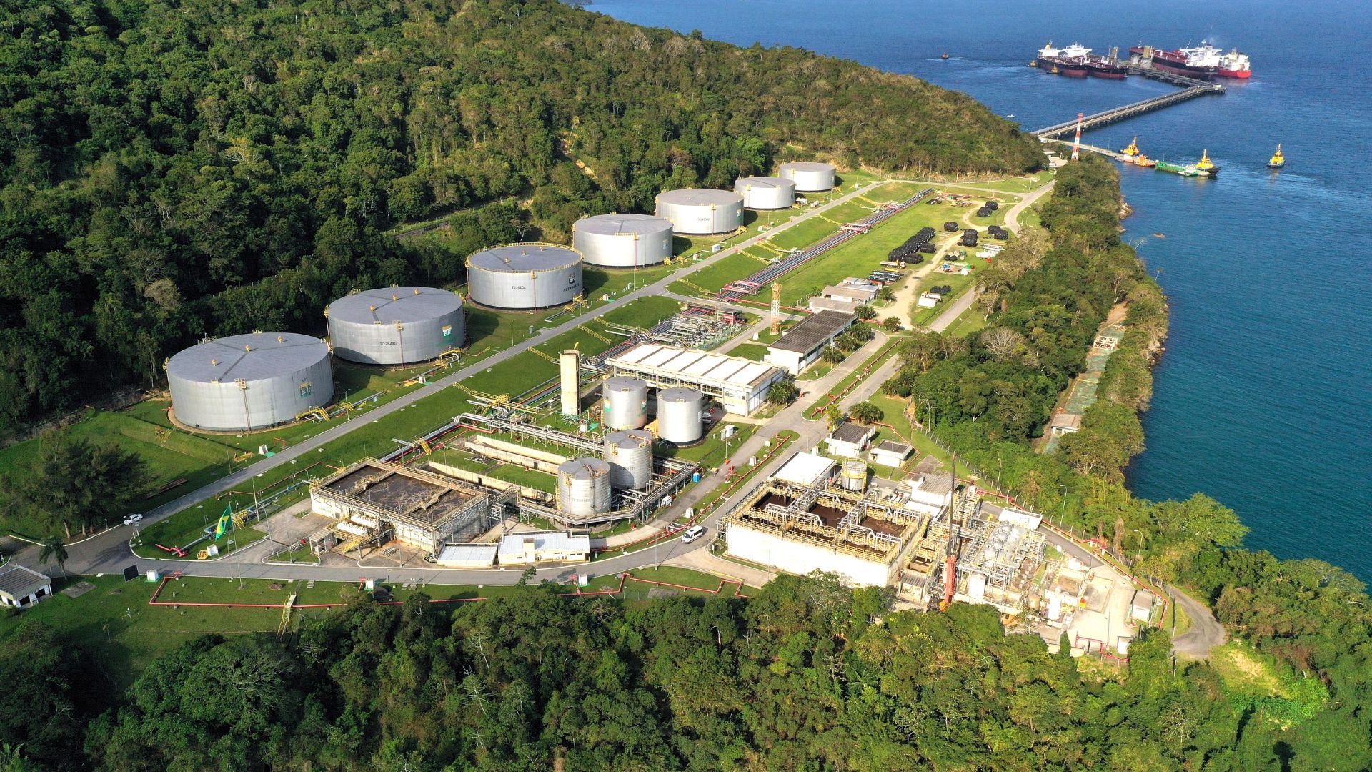 Terminal estratégico da Transpetro à beira-mar com tanques de armazenamento de petróleo e derivados, dutos interligados e navios operando em área costeira no Brasil.