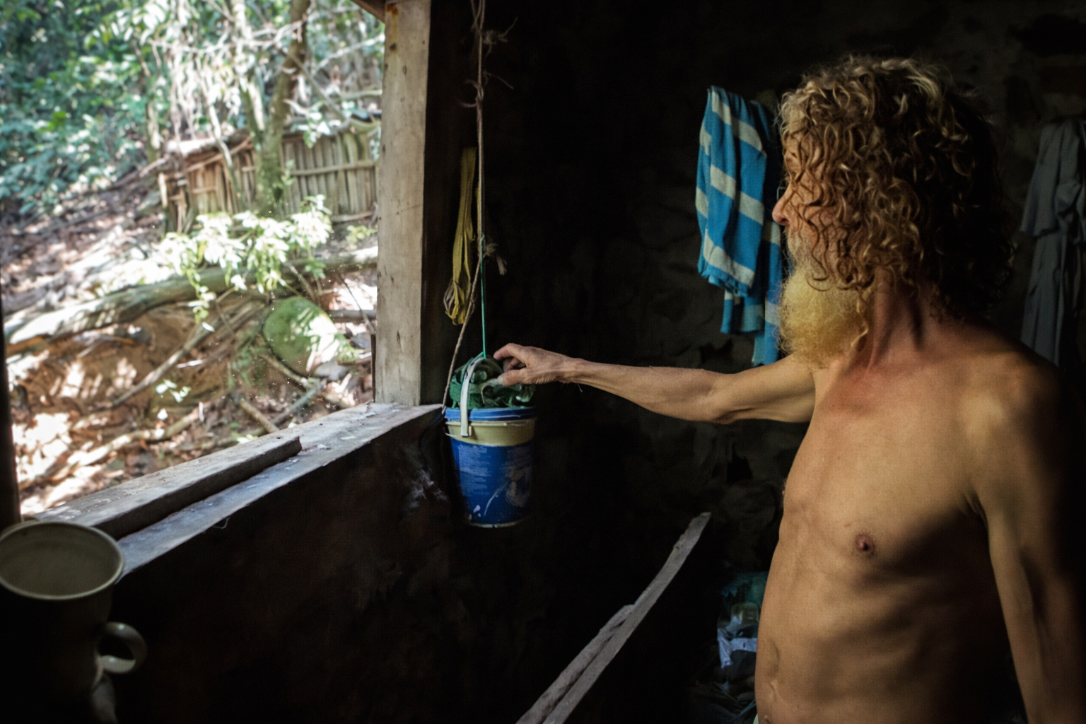 Homem vive afastado da cidade dentro de casa rústica na floresta da Pedra Branca, realizando tarefas diárias com luz natural e sem uso de tecnologia.