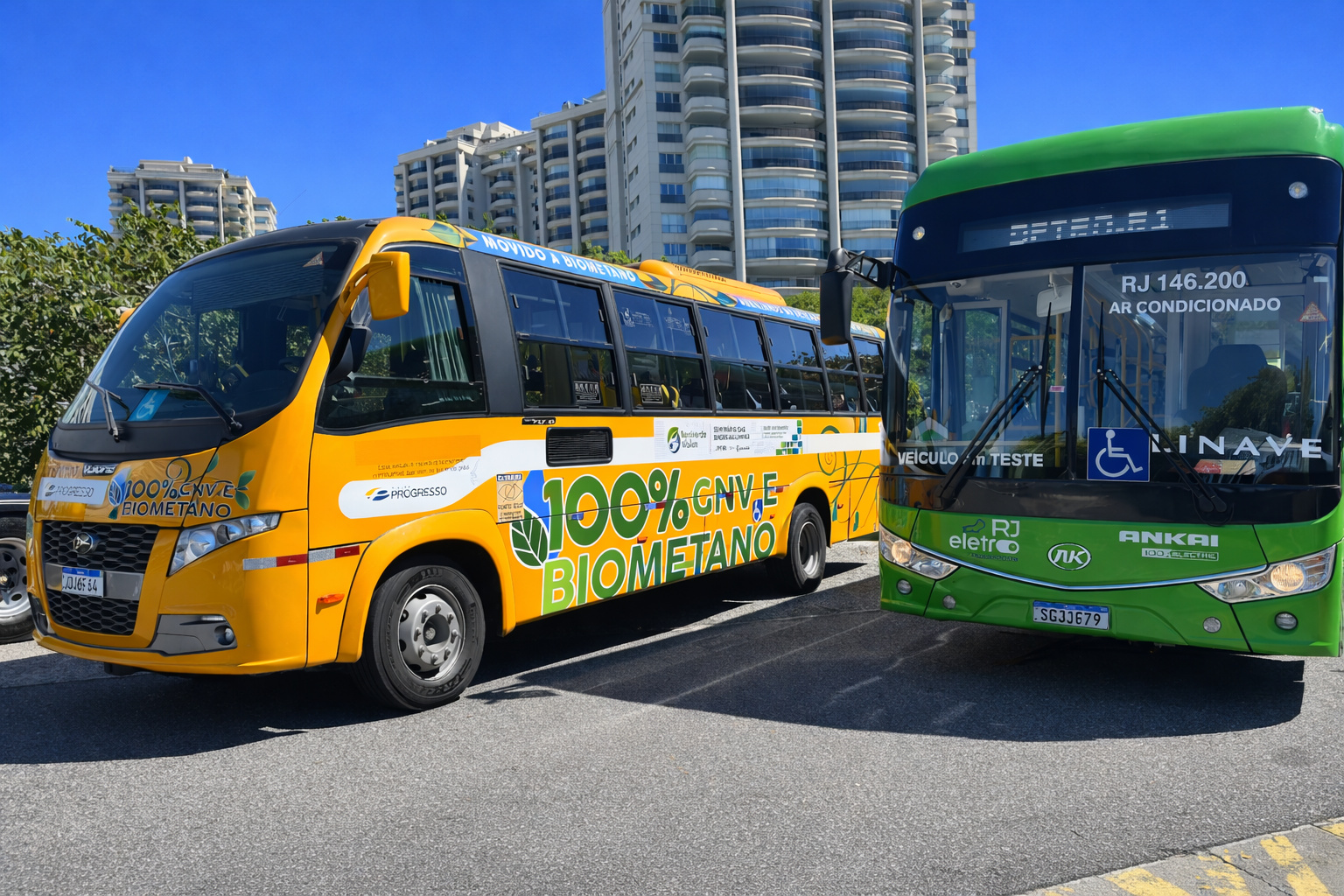 Ônibus sustentável movido a biometano e ônibus elétrico estacionados em via urbana sob céu azul ao meio-dia.