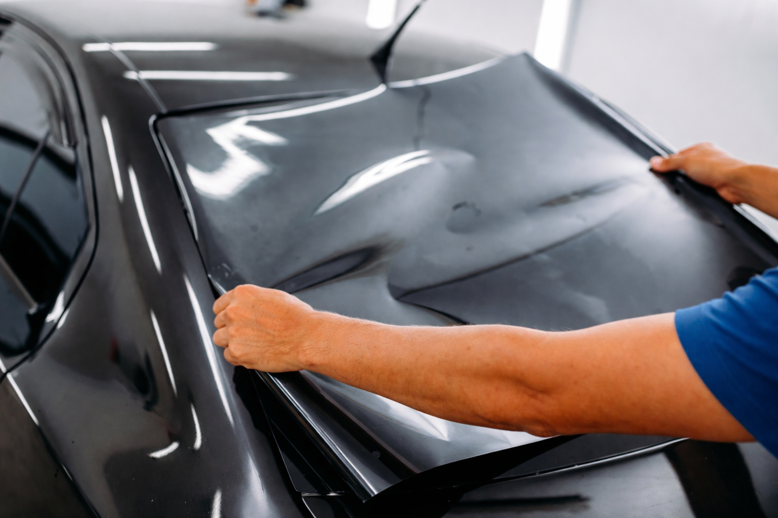 Profissional aplicando película automotiva no vidro traseiro de um carro preto, com braços morenos, em ambiente interno bem iluminado.