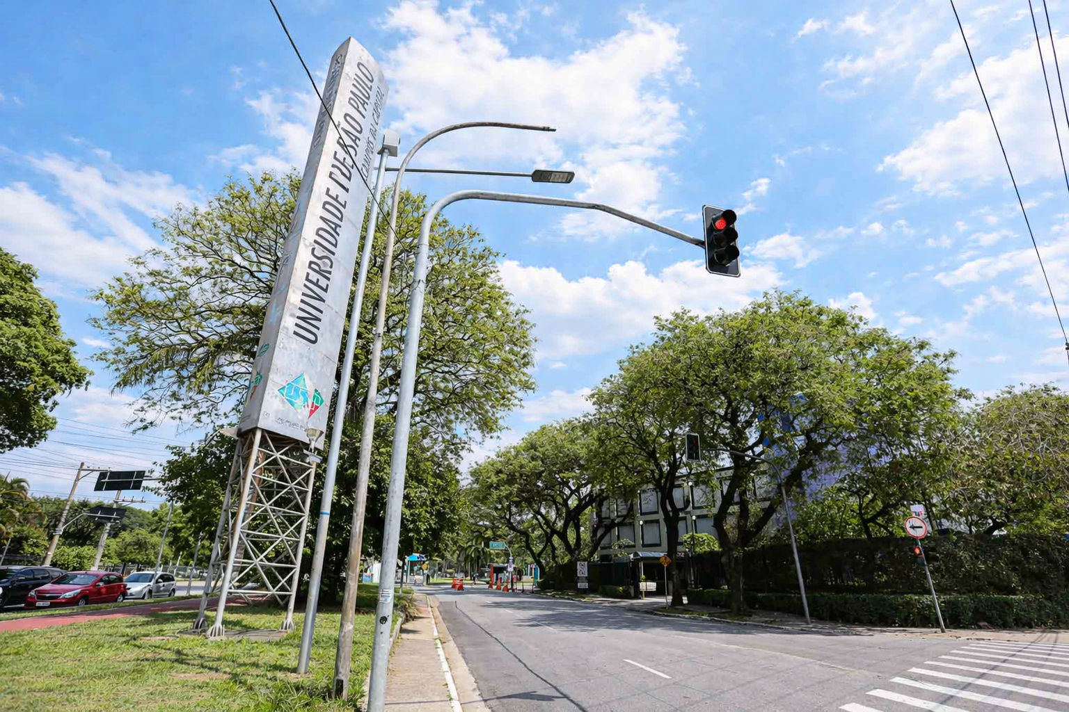 Entrada da Universidade de São Paulo com placa institucional, semáforo e árvores sob céu parcialmente nublado durante a tarde.