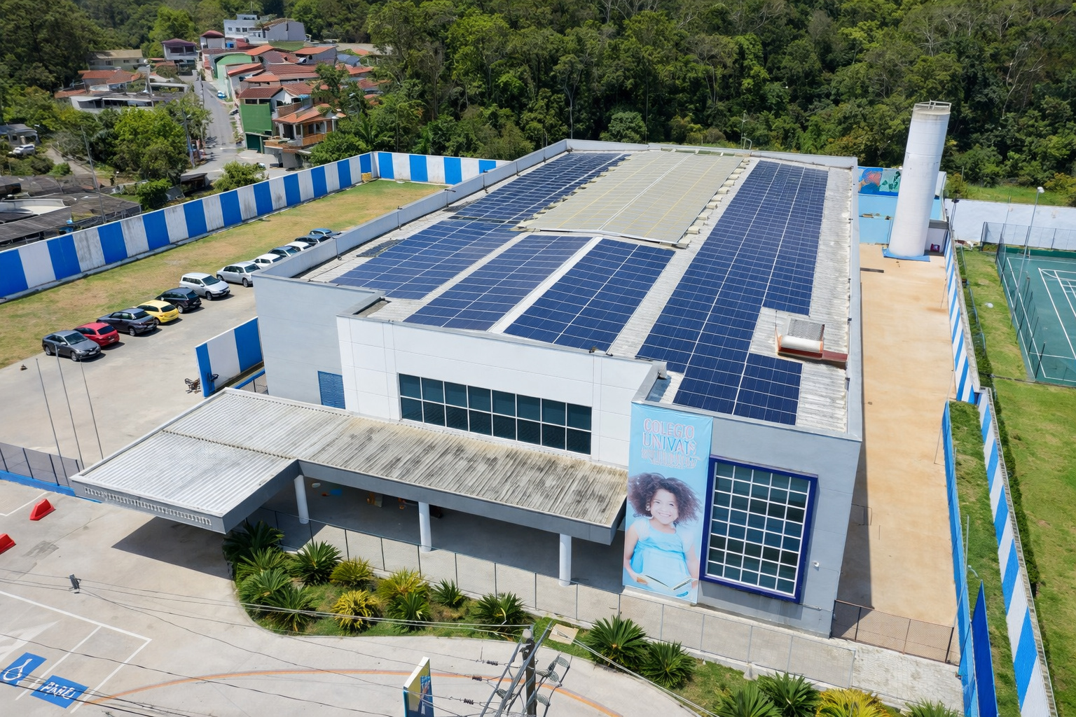 Vista aérea de prédio com painéis solares no telhado ao meio-dia, cercado por área verde, estacionamento e quadra esportiva.