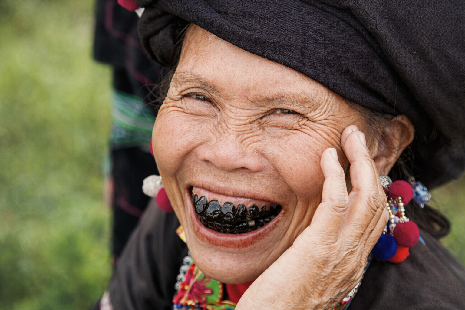 Idosa vietnamita sorrindo com dentes pintados de preto, usando traje tradicional e acessórios coloridos típicos da cultura local.