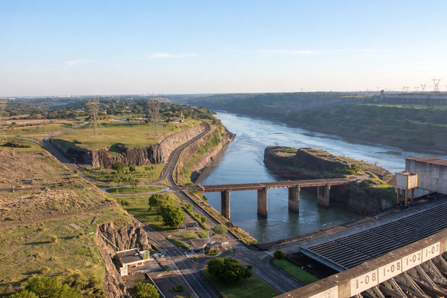 Vista aérea da Usina Hidrelétrica de Itaipu no Rio Paraná, mostrando a barragem, ponte sobre o rio, estradas de acesso e parte do complexo energético entre Brasil e Paraguai.